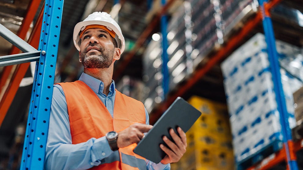 Gestionnaire d’entrepôt portant un gilet réfléchissant et un casque de sécurité à l’aide d’une tablette numérique, contrôlant les stocks et planifiant les tâches dans l’entrepôt