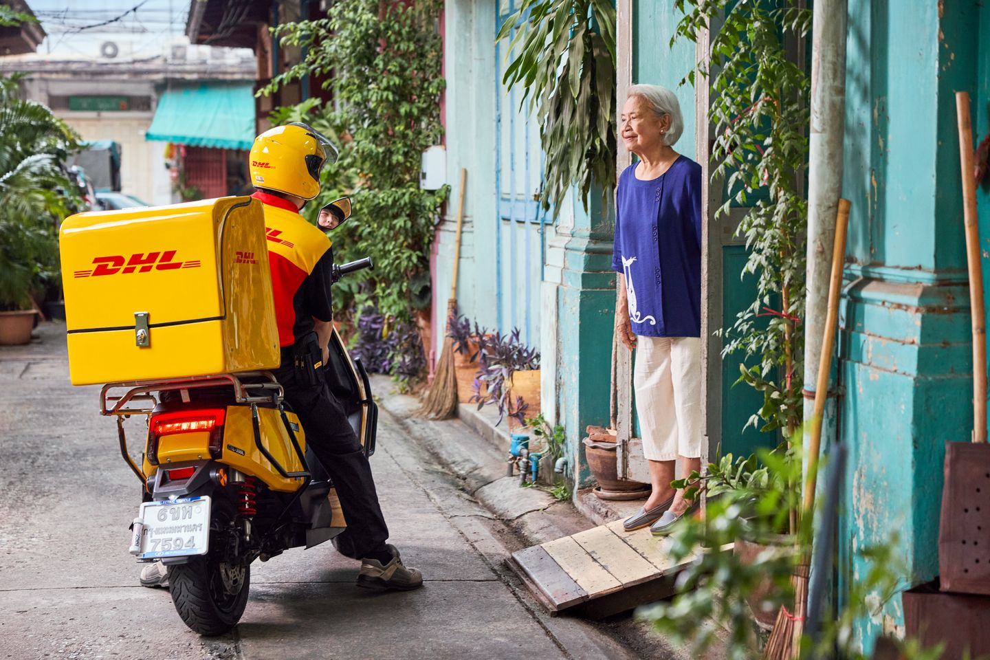 An elderly woman kindly opens the front door for a male DHL employee, who is delivering a parcel with his electric motorcycle.