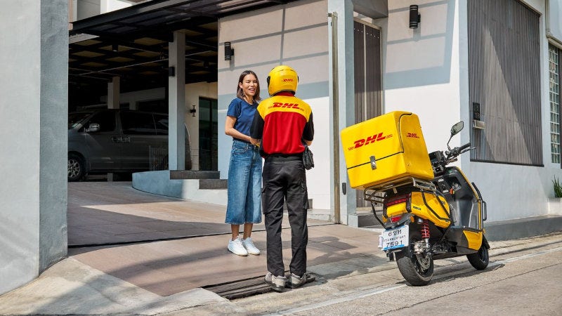 A male DHL employee delivering a parcel to a smiling female customer standing at an open gate at her modern house.
