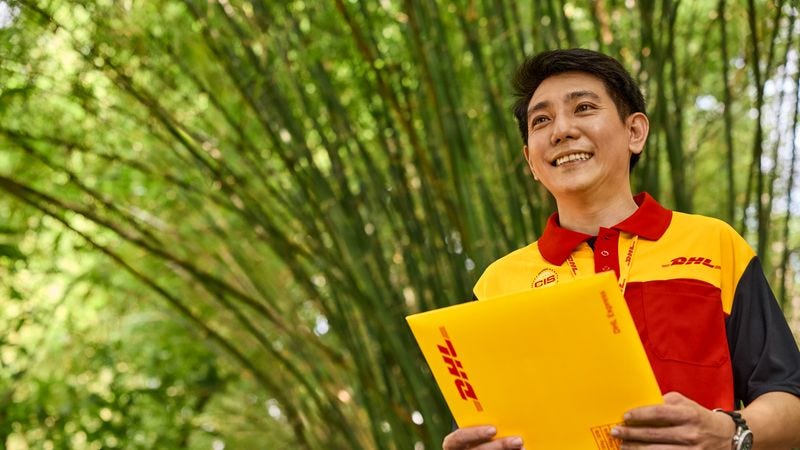 A male DHL employee holding an express envelope while standing in front of greenery.