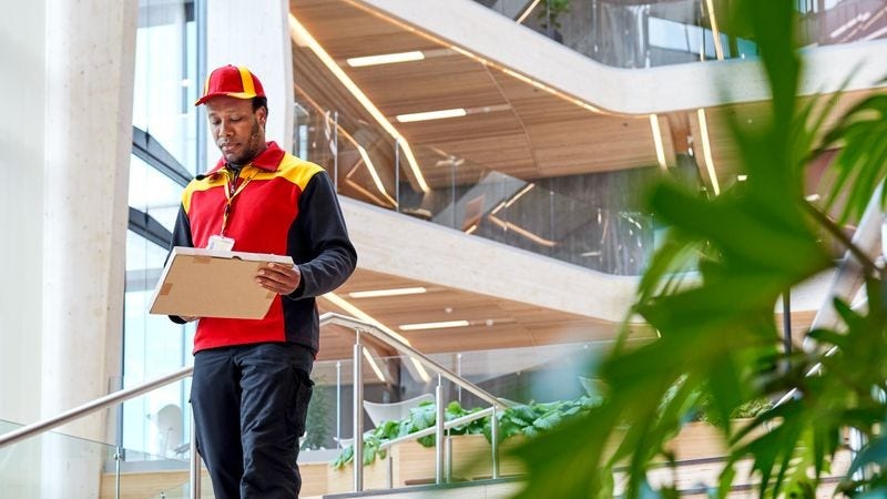 A male DHL employee is walking through a wooden office building with a parcel in his hands.