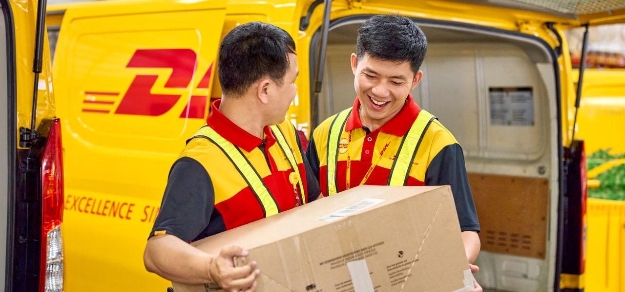 Two male DHL employees load a large parcel together into an electric DHL van.