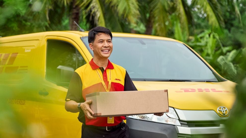 A male DHL employee stands smiling in front of a DHL van and holds a parcel in his hand.
