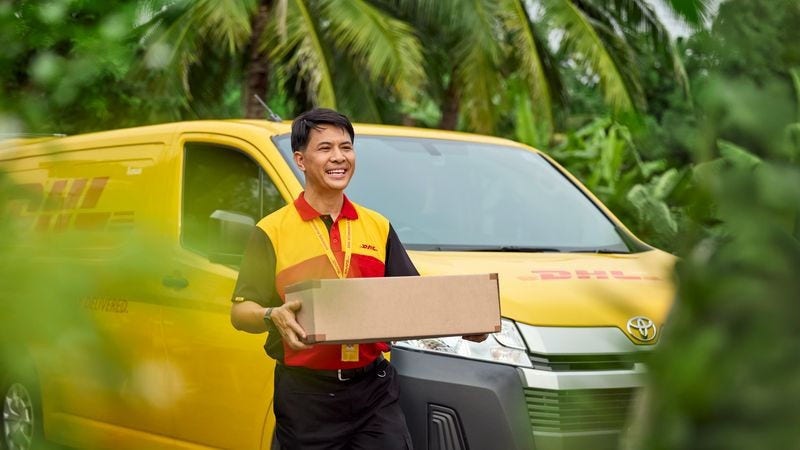A male DHL employee stands smiling in front of a DHL van and holds a parcel in his hand.