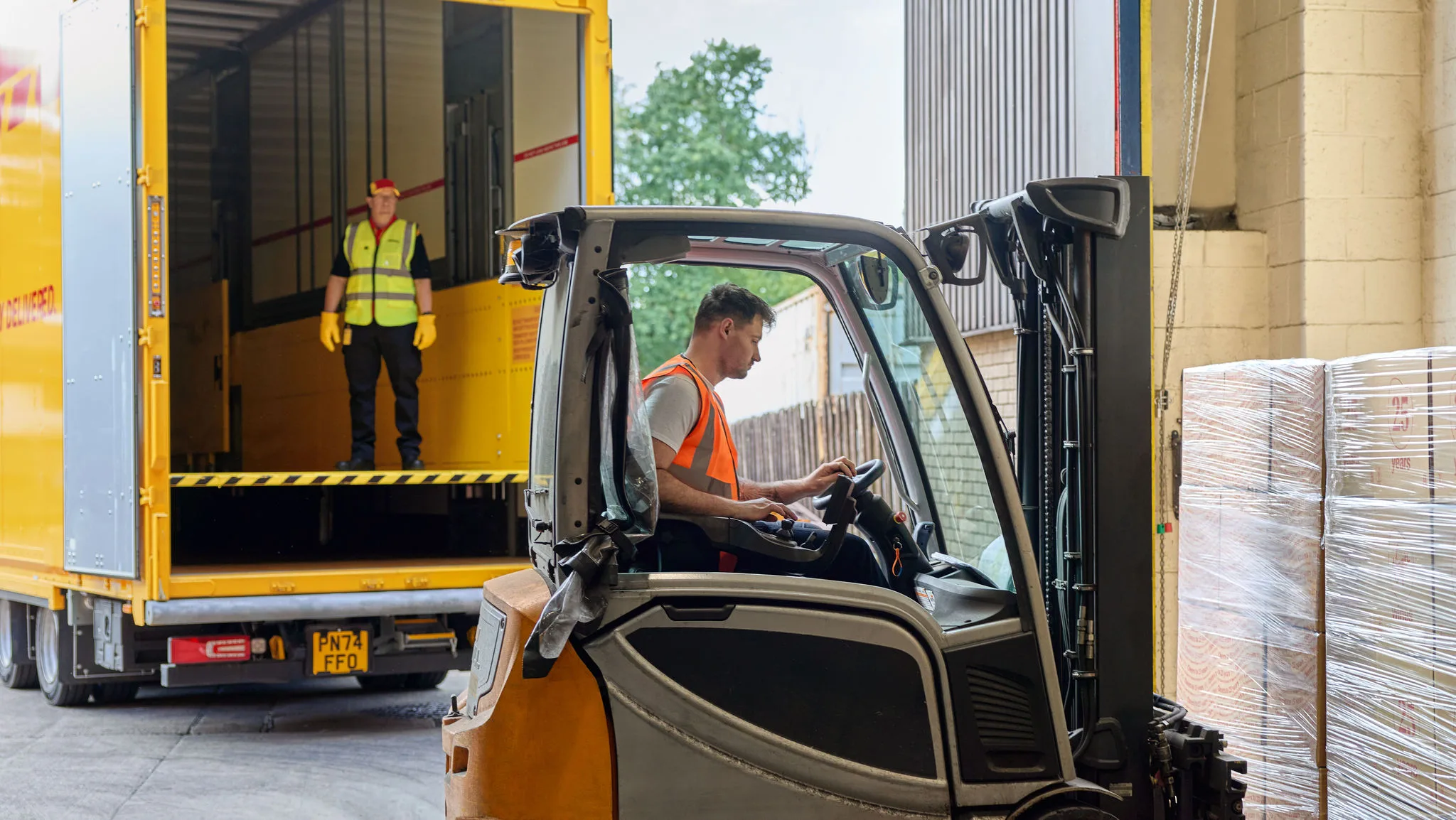 A male Virgin Wines employee loads a pallet with a forklift. A male DHL employee is standing inside the cargo area of a DHL truck in the background.