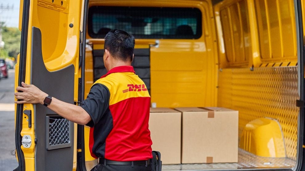 A male DHL employee loads brown parcels into the cargo area of ​​his electric DHL van.