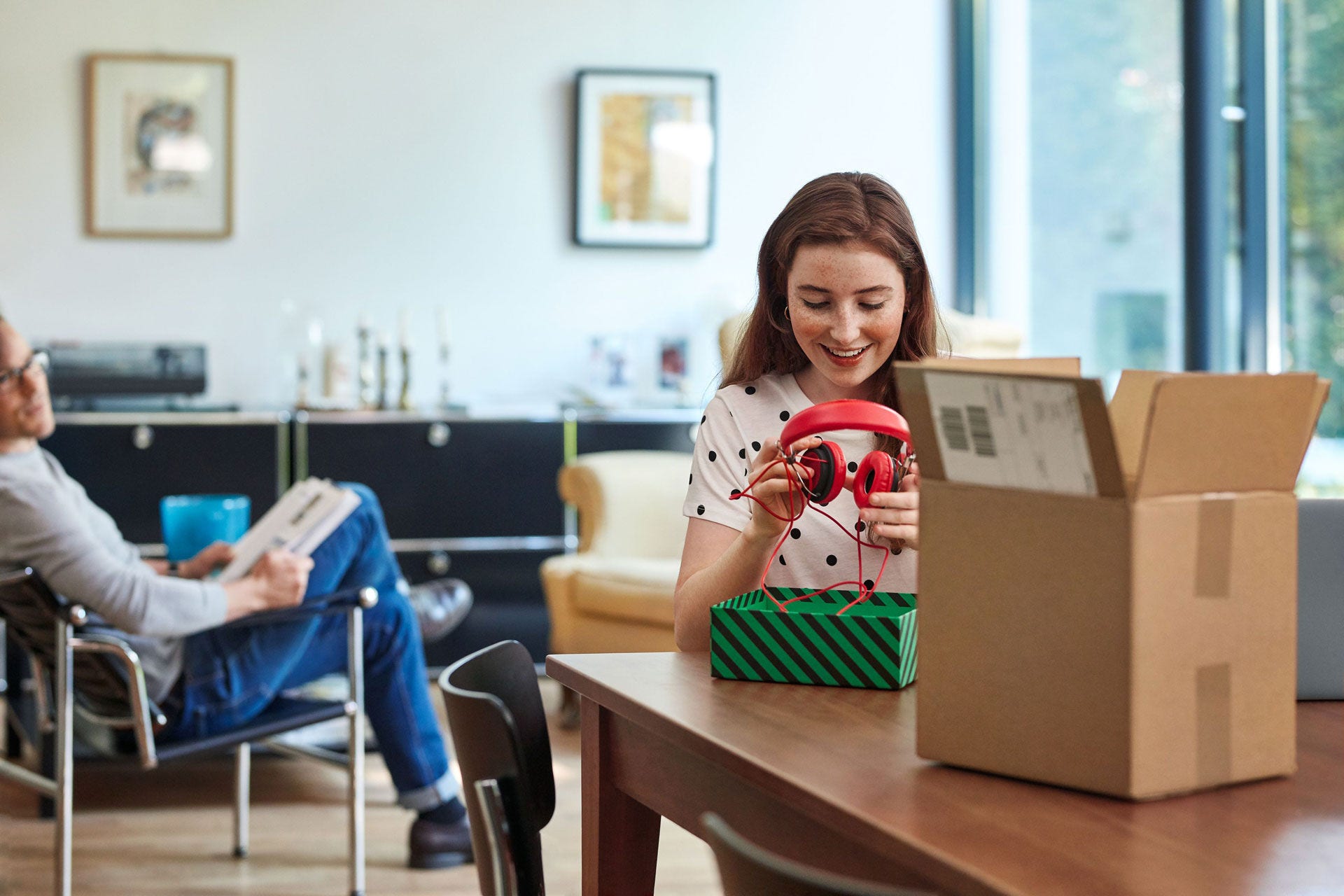 Girl holds headphones, opened parcel in the foreground, father in the background