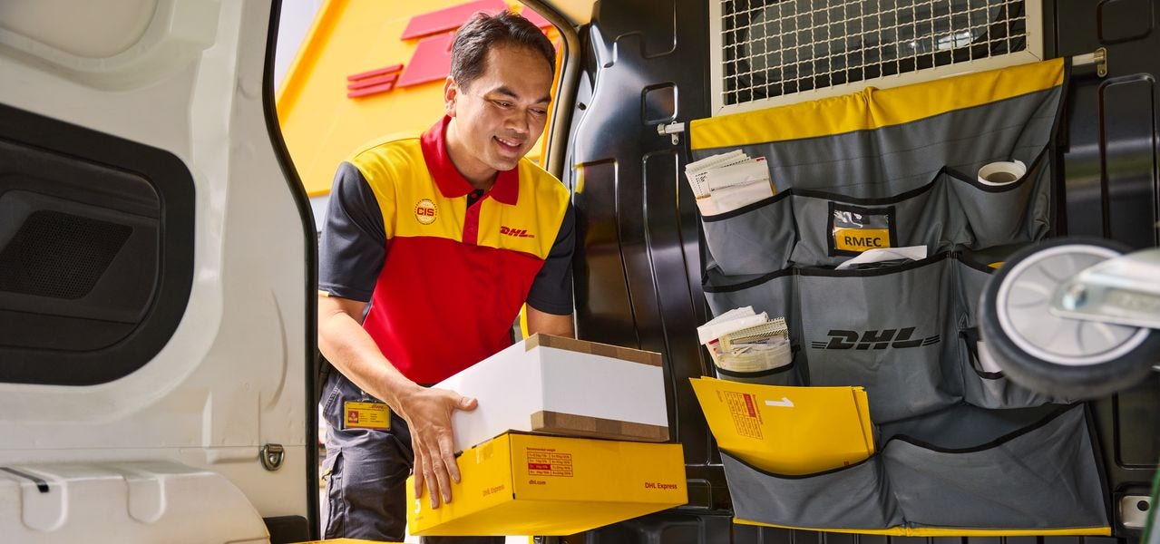 A male DHL employee loads several parcels into the loading zone of his electric DHL van.