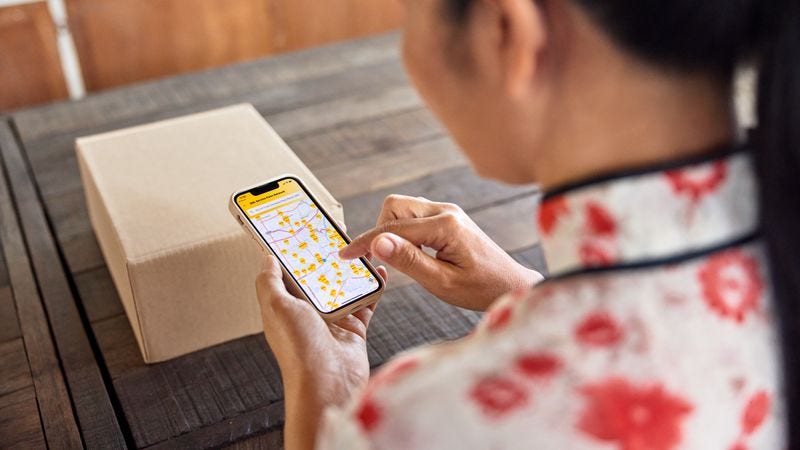 A female customer searches for the nearest DHL service point on her smartphone. Next to her on the table, a parcel is placed