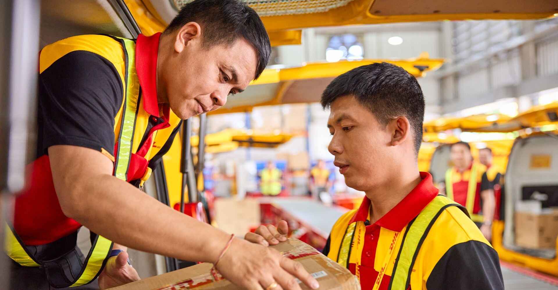 Two male DHL employees load a large parcel together into an electric DHL van.