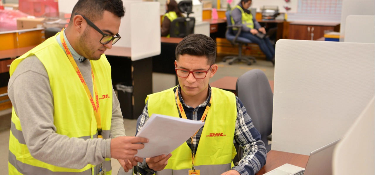 DHL 仓储办公区，两名身着反光安全背心的工作人员一同查看纸质单据，核对物流文件信息
