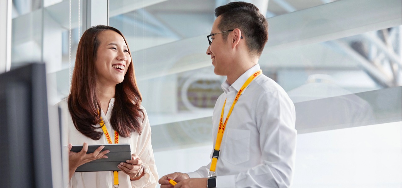 明亮的玻璃办公区，两位佩戴 DHL 黄色挂绳的员工手持平板，面带笑容交流物流业务方案