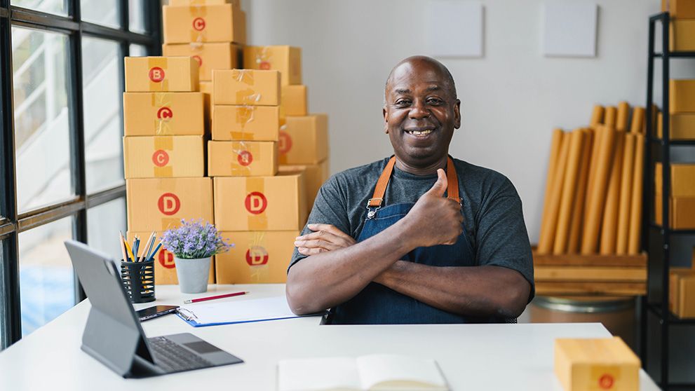Happy small business owner in apron giving thumbs up in modern office with cardboard boxes, laptop, and notepad.