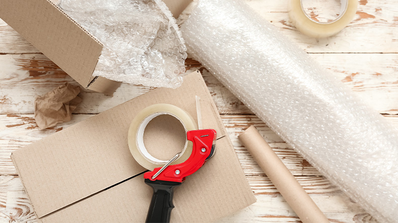 Packing tape dispenser, cardboard boxes and roll of bubble wrap on wooden background