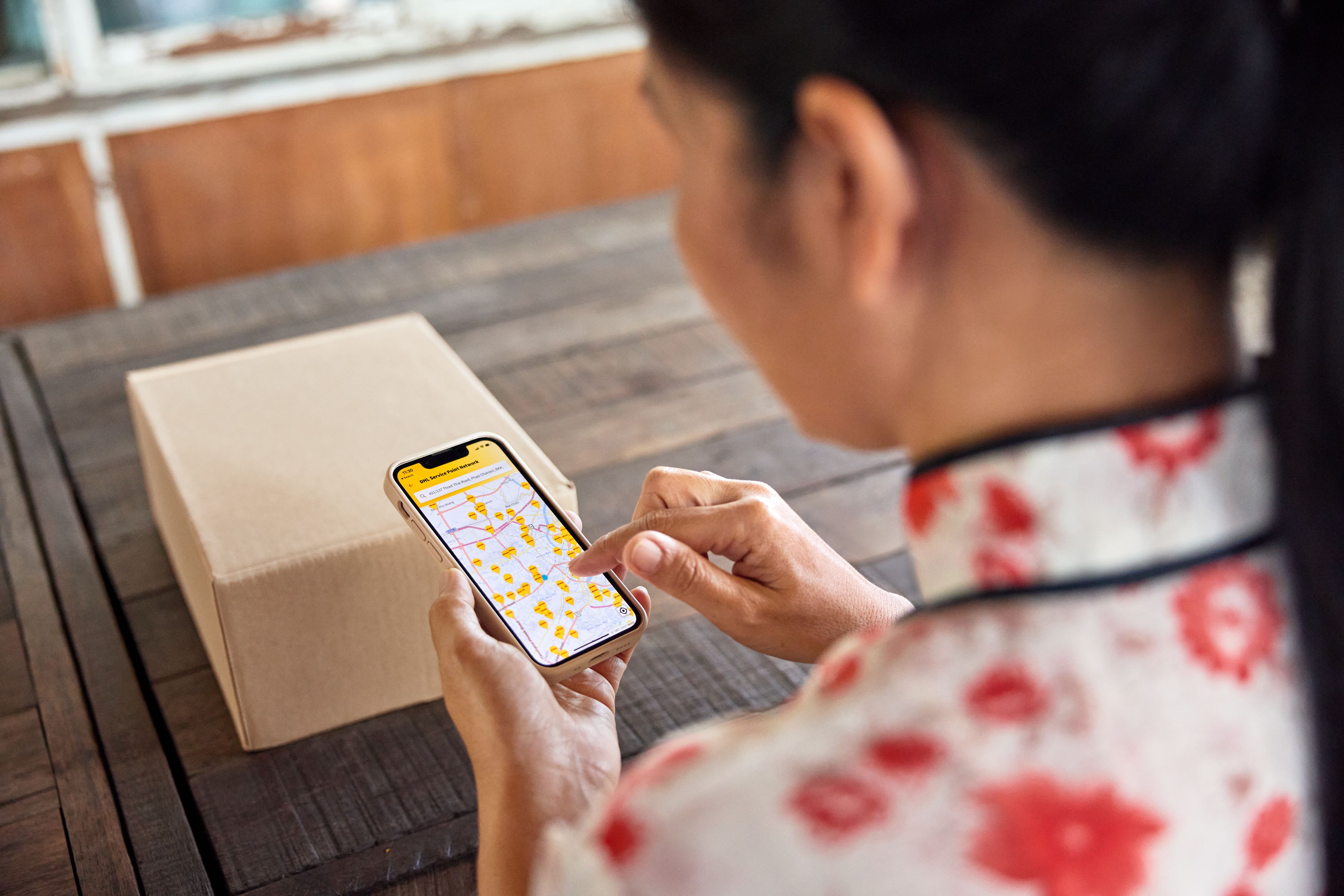 A female customer sits at the dining table in her traditional Thai home and searches for the nearest DHL service point on her smartphone. Next to her on the table, a parcel is placed.
