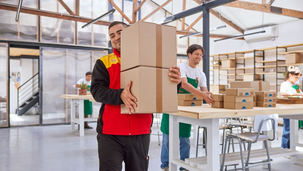 DHLの従業員が輸出用の小包を2つ抱え、その背後で複数の作業員が働いている。