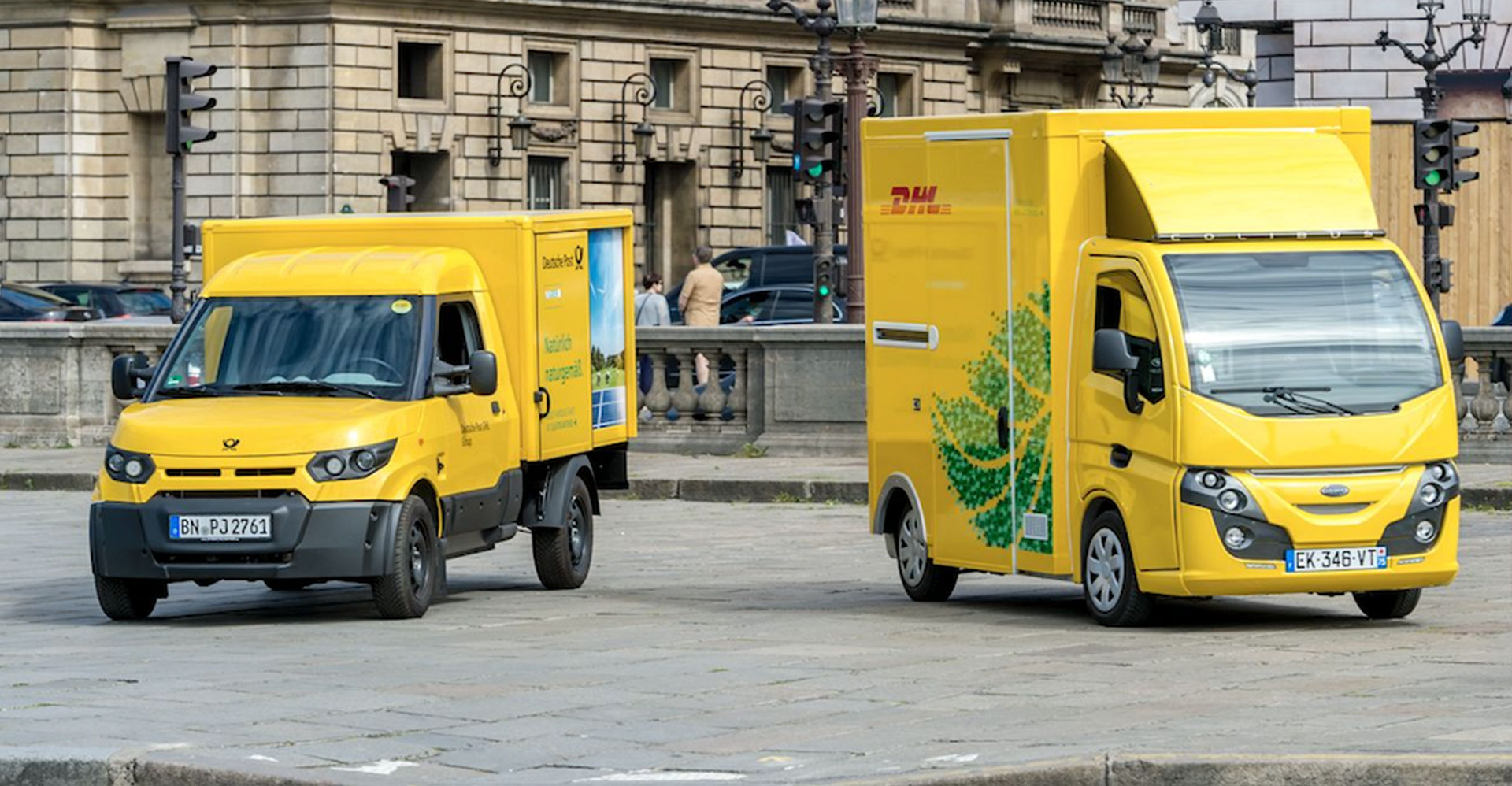 Two DHL GoGreen trucks using electric and hybrid vehicle technology parked in front of a building.