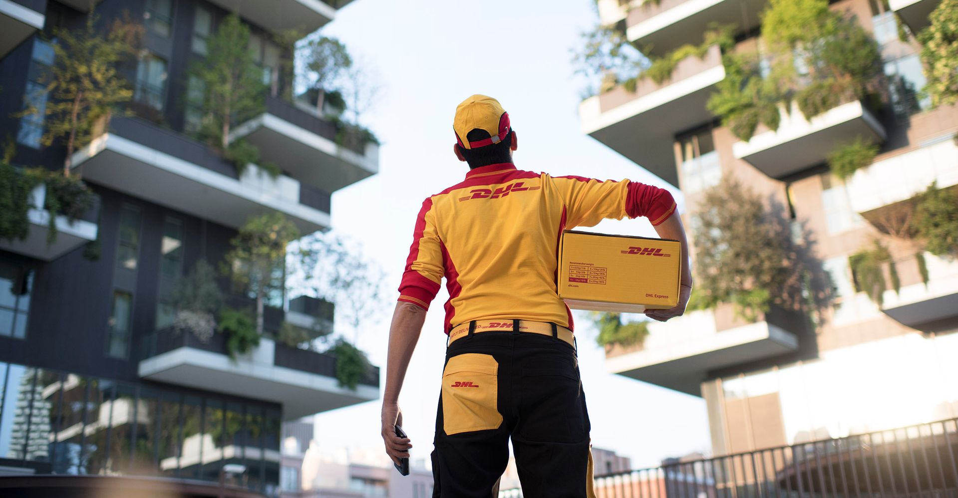 ภาพของพนักงานชาย DHL ถือกล่อง DHL และมองขึ้นไปที่อาคาร