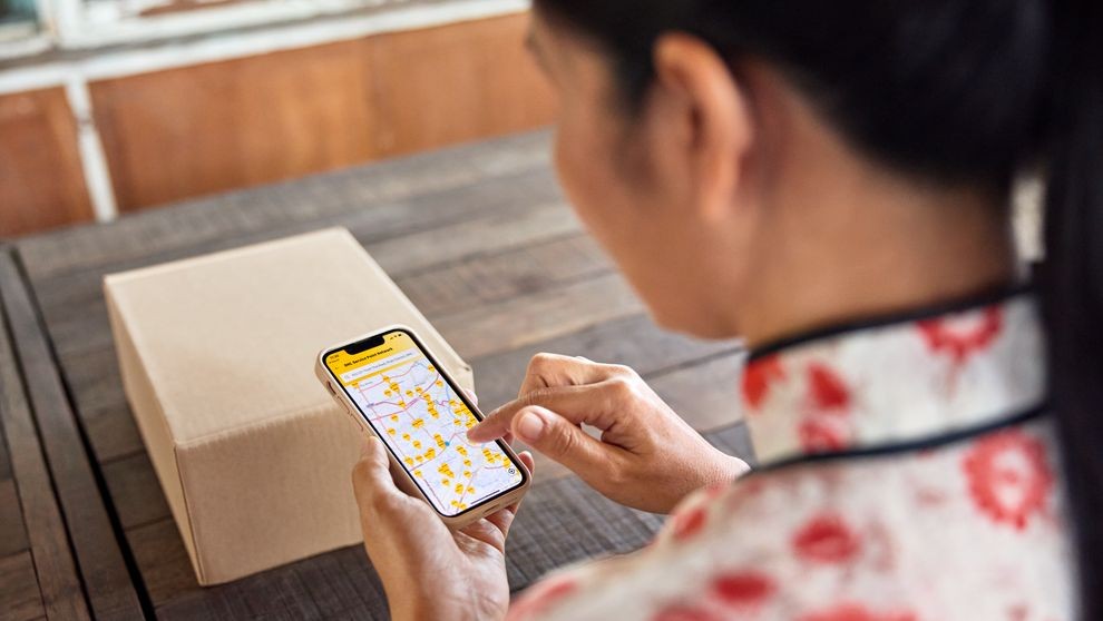 ลูกค้าหญิงจากประเทศไทยกําลังค้นหาจุดบริการ DHL ที่ใกล้ที่สุดบนสมาร์ทโฟนเพื่อส่งพัสดุของเธอ
