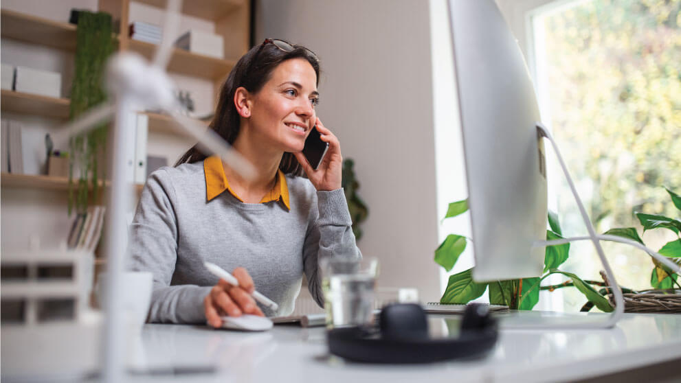 woman on her cell phone while on her computer at a desk 