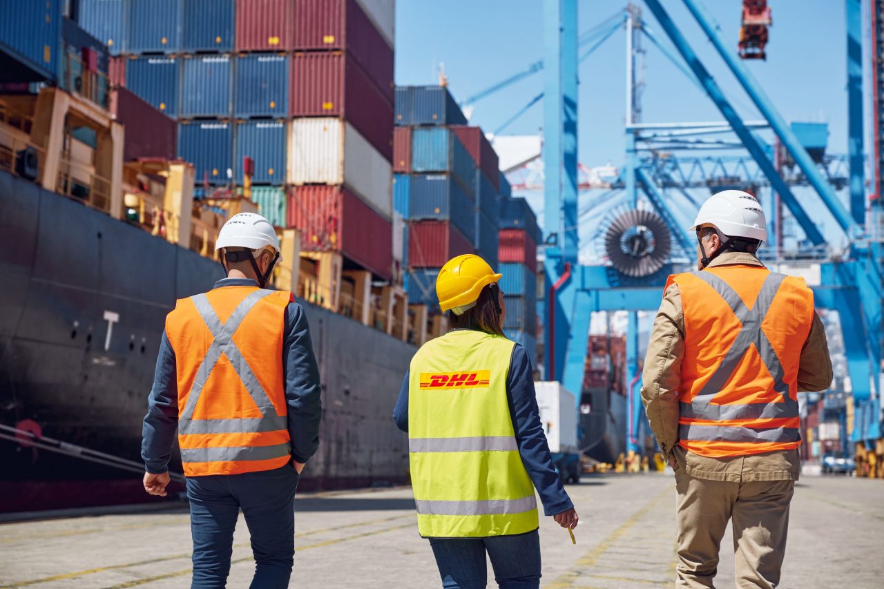 Free trade zone workers walk through an active port with shipping containers being loaded onto a ship. 