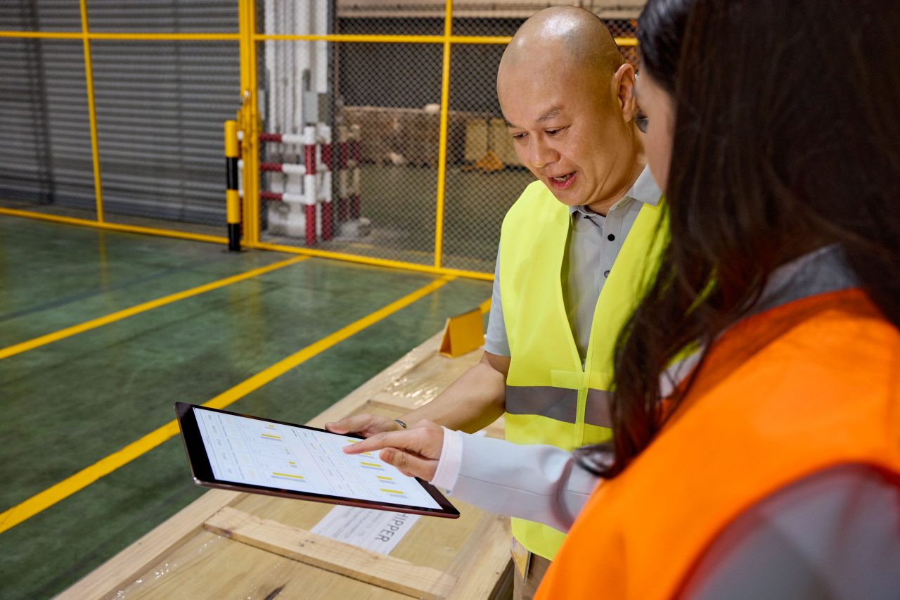 Two employees wearing high-visibility safety vests review shipping paperwork on a clipboard and tablet while standing beside a wooden pallet in a warehouse setting.