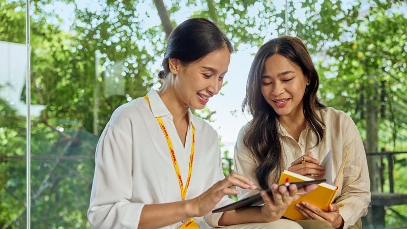 Two female DHL employees sit on a bench in a sustainable office, looking at a tablet together.