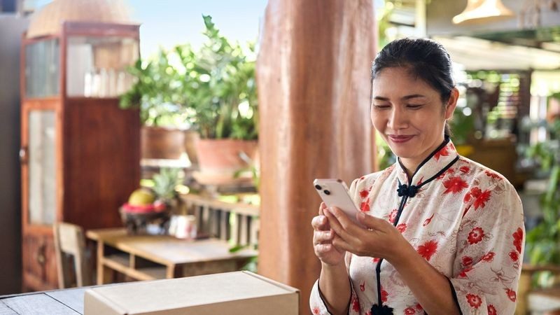 A female business owner uses social media selling platforms on her smartphone. A parcel sits on the table in front of her.