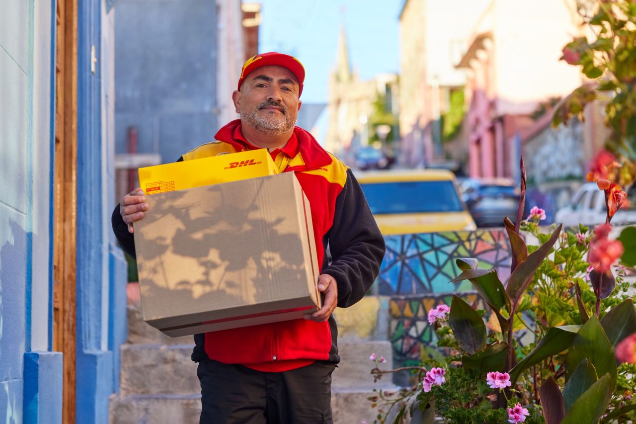A male DHL employee is carrying several parcels for delivery. His parked electric DHL van is visible in the background.