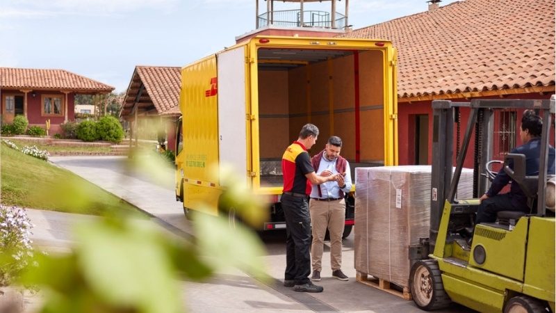 Bir erkek bağ sahibi, DHL'de erkek bir çalışanın tarayıcısını imzalamaktadır. DHL tarafından teslim edilen bir paletin yanında duruyorlar.
