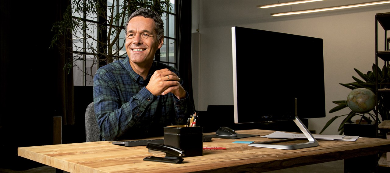 Middle-aged man wearing plaid shirt smiles sitting at his computer in a large room